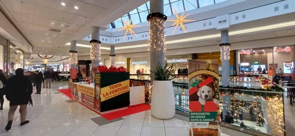 Centro Commerciale Le Brentelle decorato per Natale: stand concorso benefico 'Tenta la fortuna! Aiuta il canile di Rubano' su tappeto rosso. Colonne con tende di luci dorate, stelle luminose sospese. Cartello con cucciolo in pacco regalo 'Impacchetta un sogno aiuta il canile'. Clienti che passeggiano, negozi e decorazioni natalizie sullo sfondo