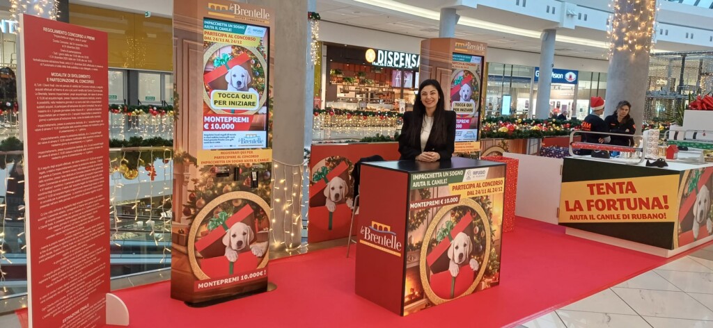 Stand completo concorso benefico Le Brentelle con hostess al banco informazioni. Cartelli 'Tocca qui per iniziare', 'Montepremi €10.000', totem con cucciolo in pacco regalo, regolamento concorso su pannello rosso a sinistra, cartello 'Tenta la fortuna!' a destra. Sullo sfondo: Dispensa Emilia e decorazioni natalizie del centro.