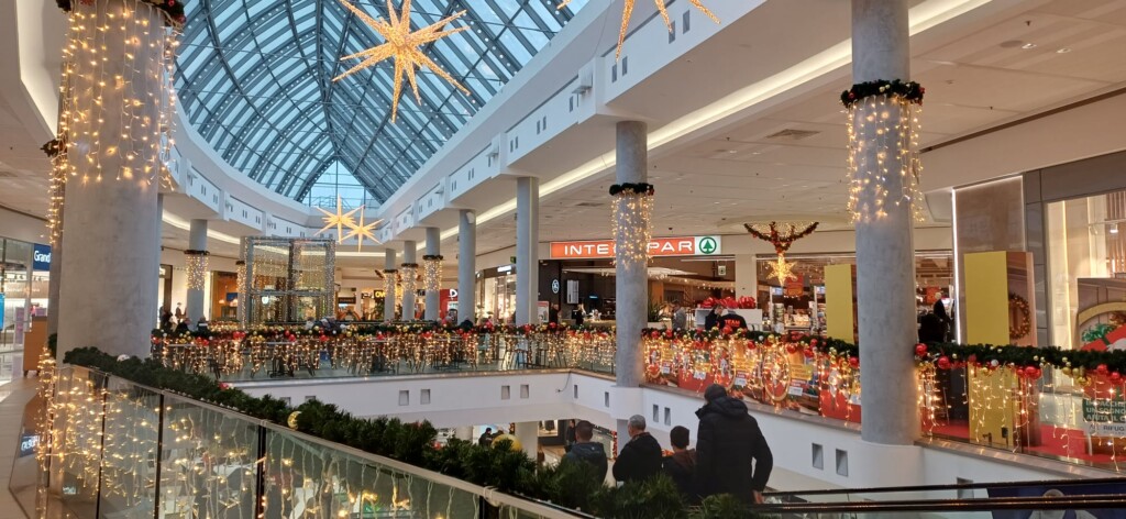 Vista panoramica Centro Commerciale Le Brentelle con decorazioni natalizie: soffitto vetrato a lucernario, stelle dorate luminose sospese, colonne con tende di luci, ringhiere decorate con ghirlande luminose rosse e dorate su più piani. Insegna Interspar visibile. Clienti sui piani. Architettura moderna luminosa